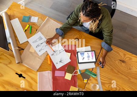 Overhead of biracial male fashion designer sitting at desk using tablet and sketches at studio. Business, work, creativity, communication, fashion and Stock Photo