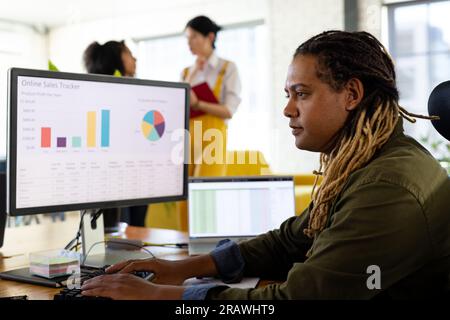 Biracial casual businessman with dreadlocks sitting at desk working at computer in office. Work, business, communication and casual office, unaltered. Stock Photo