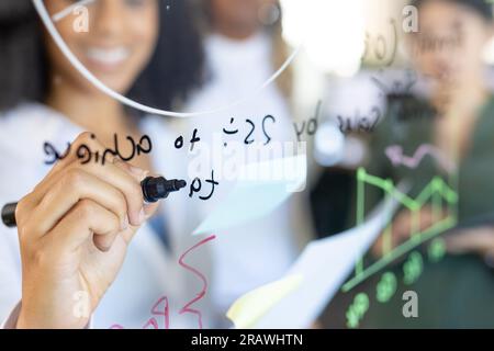 Smiling biracial casual businesswoman brainstorming with colleagues making notes on glass wall. Teamwork, work, business, brainstorm, meeting and casu Stock Photo