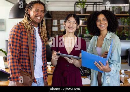 Portrait of happy diverse casual business colleagues standing in sunny office with tablet and folder. Teamwork, work, business and casual office, unal Stock Photo