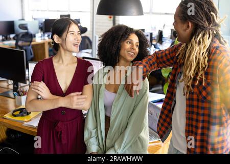Three happy diverse casual business colleagues standing in sunny office relaxing and talking. Teamwork, work, business, friendship and casual office, Stock Photo