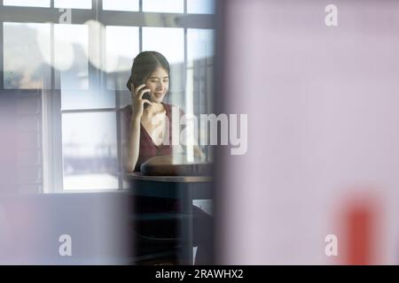 Happy asian casual businesswoman sitting at desk talking on smartphone in office, copy space. Work, business, communication and casual office, unalter Stock Photo