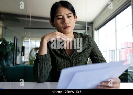 Concerned asian casual businesswoman at desk in office having video call, looking at documents. Work, business, communication and casual office, unalt Stock Photo