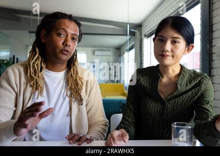 Two diverse casual business colleagues sitting at desk having video call in office. Teamwork, work, business, communication and casual office, unalter Stock Photo