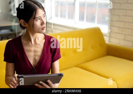 Thoughtful asian casual businesswoman with tablet sitting on sofa in office looking away, copy space. Work, business, communication and casual office, Stock Photo