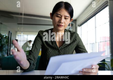 Concerned asian casual businesswoman at desk in office having video call, looking at documents. Work, business, communication and casual office, unalt Stock Photo