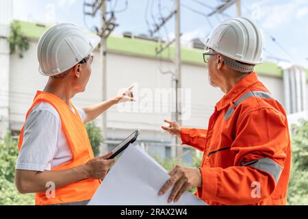 Back view of 2 male engineers or foremen in orange jumpsuit, safety vest and white hardhat, one holding a computer tablet and the other holding a buil Stock Photo