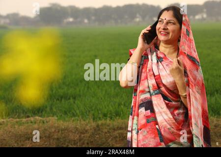 Happy rural Indian woman standing in a mustard agricultural field and surfing through the mobile phone in her hand. Stock Photo