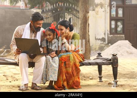 Happy Indian Rural family of Mother, father and daughter sitting together smiling on cot outside their home in front yard using Laptop. Stock Photo