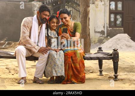 Happy Indian Rural family of Mother, father and daughter sitting together smiling on cot outside their home in front yard using mobile phone. Stock Photo