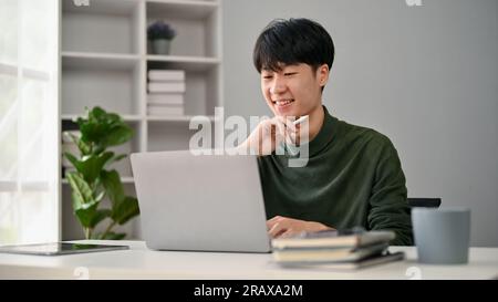 A smart and happy young Asian male office worker in casual clothes looking at his laptop screen, thinking creative ideas, planning his project, workin Stock Photo