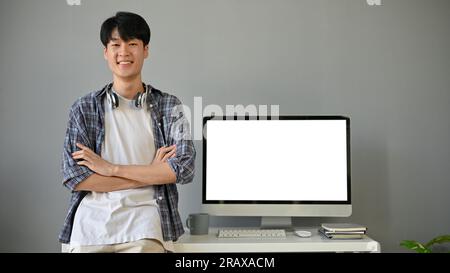 A smart and handsome young Asian male programmer in a casual clothes stands with his arms crossed in his modern office. A computer white screen mockup Stock Photo