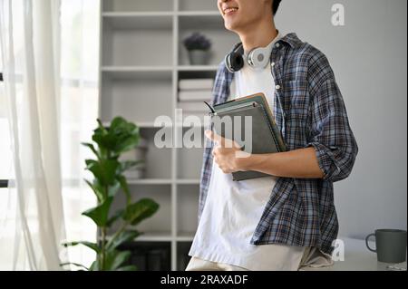 Cropped image of a smiling handsome young Asian man in a flannel shirt sitting on a table with books in his hand. Stock Photo