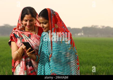 Happy rural Indian women standing in a mustard agricultural field and surfing through the mobile phone in their hand. Stock Photo