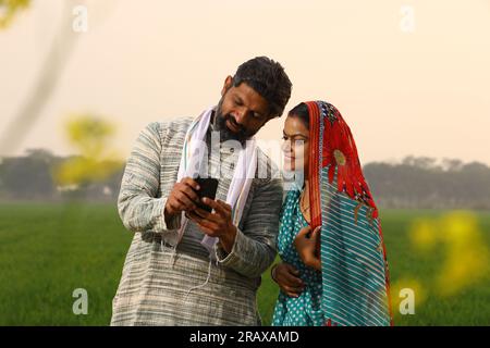 Happy rural Indian villager family standing in a mustard field husband showing his wife something on his mobile smart phone screen and laughing Stock Photo