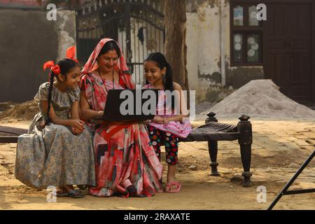 Happy Indian Rural family in village. Mother and daughters sitting together smiling on cot outside their home in front yard. Mother in beautiful saree Stock Photo