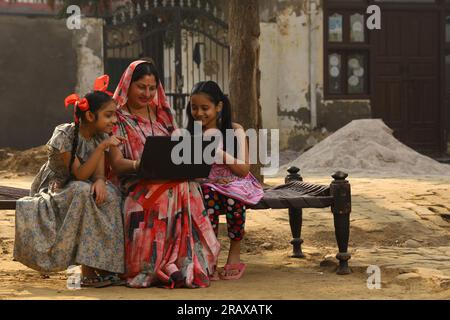 Happy Indian Rural family in village. Mother and daughters sitting together smiling on cot outside their home in front yard. Mother in beautiful saree Stock Photo