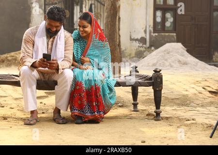 Happy Indian Rural family in village. couple sitting together outside their home front yard using mobile phone. Wife beautiful saree traditional wear Stock Photo