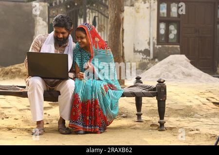 Happy Indian Rural family in village. Husband and wife sitting on cot outside their home front yard. man in kurta pajamas and wife in beautiful saree. Stock Photo