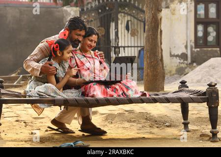 Happy Indian Rural family Mother,father,daughter sitting together on cot outside their home front yard using Laptop. Mother in beautiful saree. Indian Stock Photo