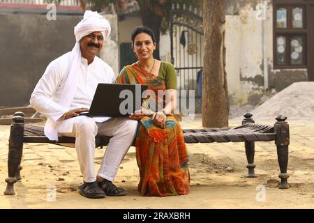 Happy Indian Rural family in village. Husband and wife sitting on cot outside their home front yard. man in kurta pajamas and wife in beautiful saree. Stock Photo