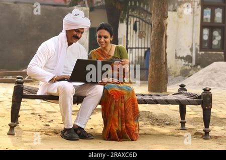 Happy Indian Rural family in village. Husband and wife sitting on cot outside their home front yard. man in kurta pajamas and wife in beautiful saree. Stock Photo