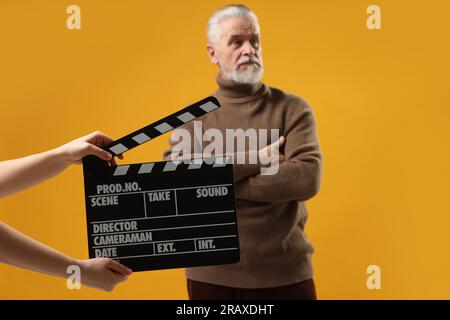 Senior actor performing while second assistant camera holding clapperboard on yellow background, selective focus. Film industry Stock Photo