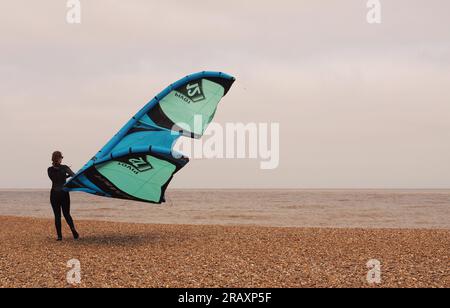 A female windsurfer standing on the shingle beach at Aldeburgh, Suffolk. UK holding onto her windsurfing sail in windy conditions Stock Photo