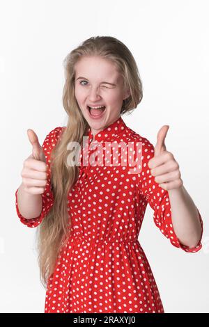 Happiness blonde young female playfully closed one eye and showing thumbs up with both hands. Portrait of smiling young adult woman dressed in summer Stock Photo