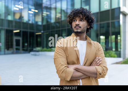 Portrait of a young Indian male programmer, developer standing with crossed arms outside the office center, seriously and confidently looking to the side. Stock Photo