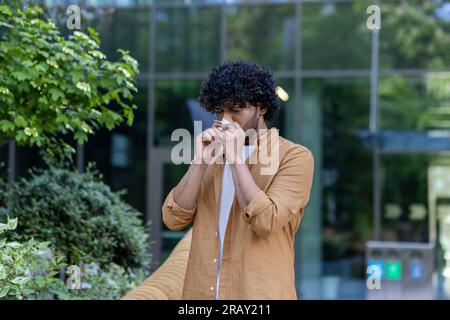 A young Indian man stands on the street and blows his nose and wipes his nose with a tissue. Suffers from runny nose, infection, seasonal allergies. Stock Photo