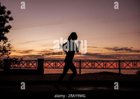 Silhouette of young woman running along the road along the sea early in the morning at the dawn of red sun. Girl is engaged in fitness to maintain sha Stock Photo