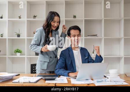 Two asian business people successful excited raised hands rejoicing with a laptop computer in office Stock Photo