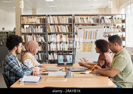Group of foreign students having online lesson with teacher while sitting in the library Stock Photo