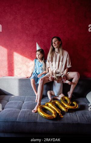 Tired young mother sits on sofa with small children. Red wall. Single parent family celebrating babys first year. Foil balloons ONE on gray couch. Lif Stock Photo