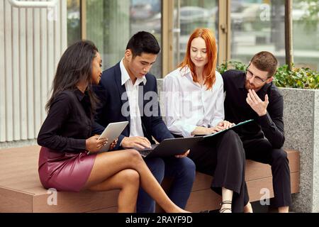 Group of employees, project leader sitting on street and working on current project together. Discussing and analytics Stock Photo