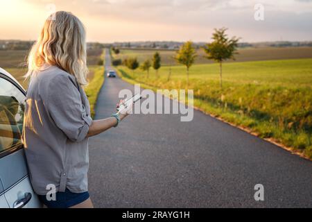 Woman using smartphone during summer road trip. Female traveler standing at car Stock Photo