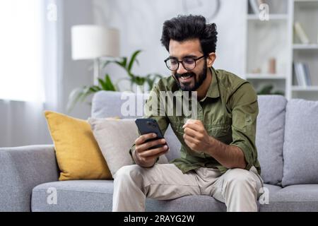 Happy and satisfied with achievement results hispanic at home reading online win notification in online application on phone, man smiling holding hand up triumph gesture sitting sofa in living room. Stock Photo