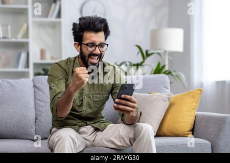 Happy and satisfied with achievement results hispanic at home reading online win notification in online application on phone, man smiling holding hand up triumph gesture sitting sofa in living room. Stock Photo