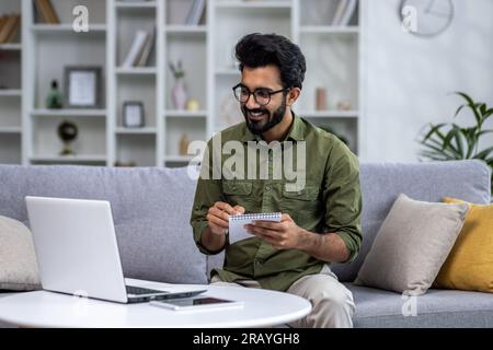 Young Indian man studying online remotely, man using laptop sitting on sofa in living room, writing outline in notebook smiling happy with virtual learning. Stock Photo