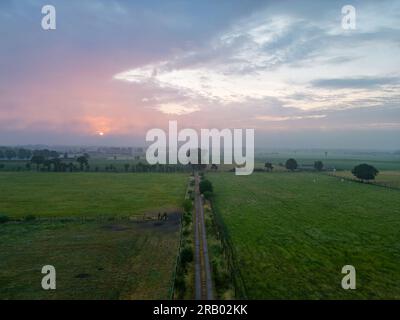 A breathtaking aerial view of a summer landscape where a winding country road leading into the fog forms the central focus. The photo, set against a dramatic sunset sky, creates an ethereal harmony between the natural beauty of the open field and the mysterious charm of the fog. A remarkable representation of nature's vastness and allure. Aerial View of Enchanting Summer Landscape: Foggy Country Road at Sunrise. High quality photo Stock Photo