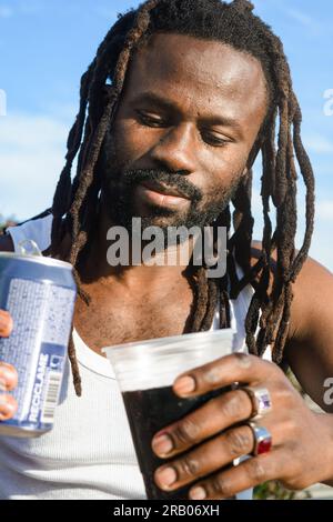 vertical portrait black man with dreadlocks and beard, happy outdoors sitting at sunset, with a cold drink in his hands Stock Photo