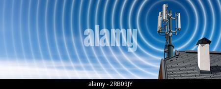 Closeup of a house roof with a telecommunications tower with antennas (aerial) and radio waves, against a clear blue sky with clouds and copy space. Stock Photo
