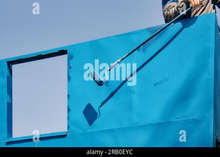 Professional painter works at height and paints empty wall surface with paint roller on summer day. Background with copy space. Resistant paint for metal. Stock Photo