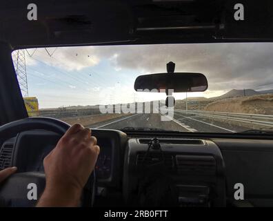 Pov point of view of man hand driving the car in scenic destination long straight roads with mountains around. Concept of transport and drive. People traveling alone. Adventure lifestyle. Vacation Stock Photo