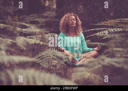 Life balance nature contact feeling serene woman doing meditation in lotus yoga posture position. Happiness and relaxation in the forest sitting with tropical leaf around. People outdoor leisure relax Stock Photo