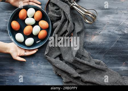 Young woman's hands placing a bowl of fresh brown and blue eggs onto a rustic wooden table. Top view. Stock Photo