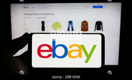 Person holding smartphone with logo of US e-commerce company eBay Inc. on screen in front of website. Focus on phone display. Stock Photo