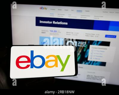 Person holding mobile phone with logo of American e-commerce company eBay Inc. on screen in front of business web page. Focus on phone display. Stock Photo