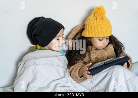 Children in warm clothes at home in cold winter time. Kids in faux fur coat, knitted hat and scarf sitting in a bed in home, warms in blanket and using tablet. Stock Photo